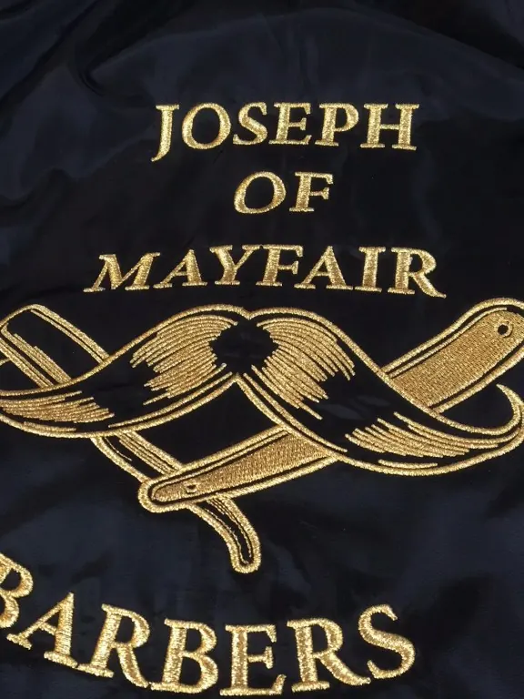 Gold thread lettering and a crest embroidered on the back of a black barber jacket.