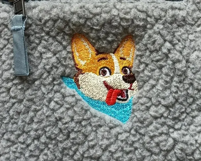 A corgi's face embroidered in colour onto a grey fleece pullover, below the chest zip.