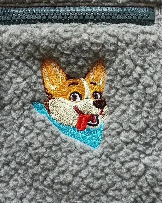 A corgi's face embroidered in colour onto a grey fleece pullover, below the chest zip.