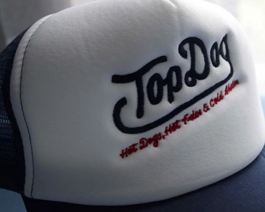 A white and navy trucker cap with a script logo embroidered on the front panel.