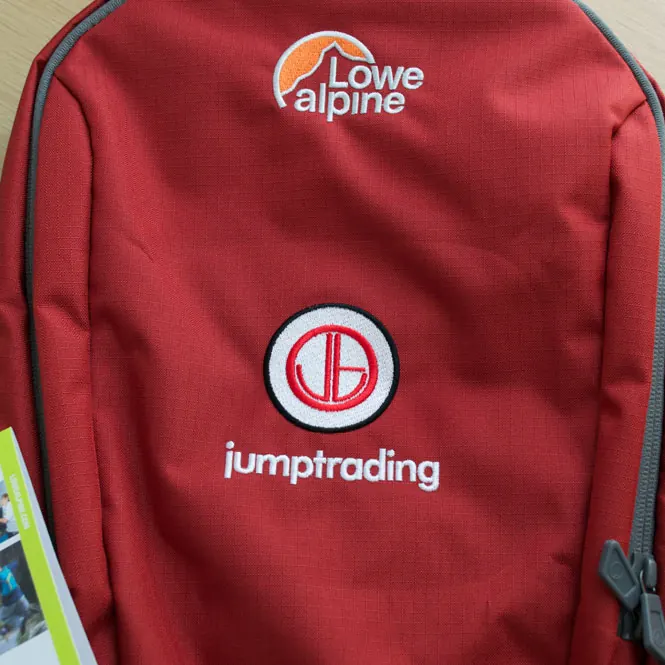 Red technical backpack with a trading company logo embroidered on the front panel.